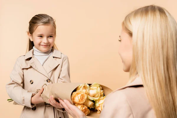Cheerful kid giving roses to mother on blurred foreground isolated on beige — Stock Photo