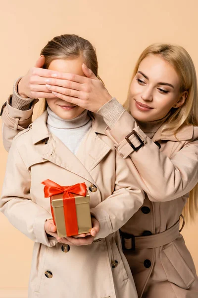 Alegre madre cubriendo los ojos de niño con caja de regalo aislado en beige - foto de stock