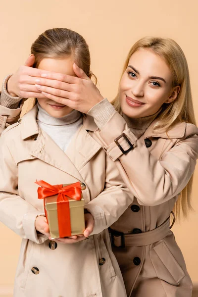 Feliz madre cubriendo los ojos de niño con caja de regalo aislado en beige - foto de stock