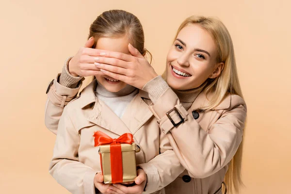 Happy mother covering eyes of child with gift box isolated on beige — Stock Photo