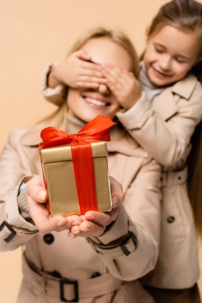 Lächelndes Kind bedeckt Augen einer glücklichen Mutter mit Geschenk isoliert auf beige — Stockfoto