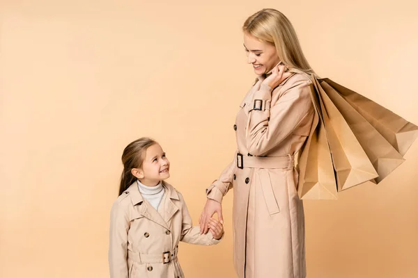 Madre feliz con bolsas de papel cogidas de la mano con el niño aislado en beige - foto de stock