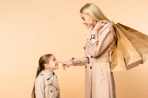 Happy mother holding paper bags and touching nose of kid isolated on beige — Stock Photo