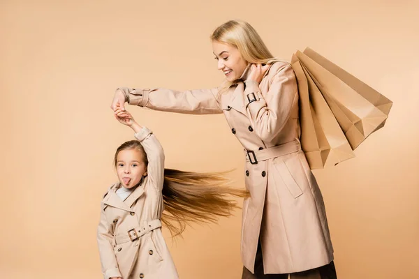 Glückliche Mutter hält Papiertüten und die Hand eines lustigen Kindes, das die Zunge isoliert auf beige ragt — Stockfoto