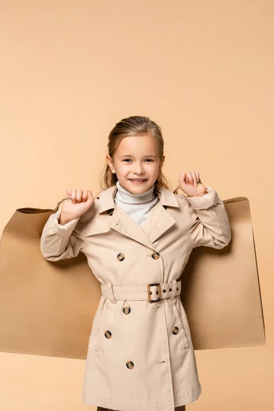 Happy kid in trench coat holding paper bags isolated on beige — Stock Photo