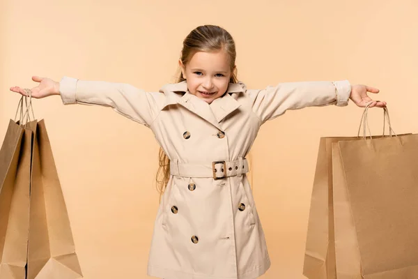 Fröhliches Kind im Trenchcoat mit Papiertüten auf Beige — Stockfoto