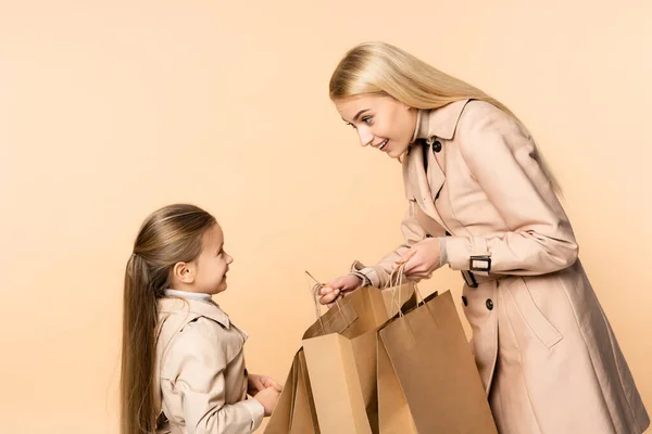 Sonriente madre sosteniendo bolsas de papel cerca alegre hija aislado en beige - foto de stock