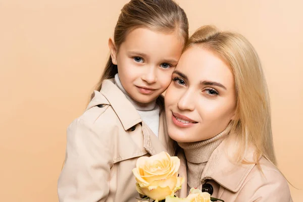 Joyful mother holding flowers and hugging daughter isolated on beige — Stock Photo