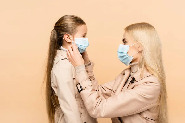 Side view of mother adjusting medical mask of daughter isolated on beige — Stock Photo