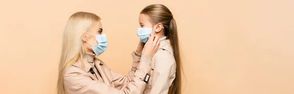 Vista laterale della madre che regola la maschera medica della figlia isolata sul beige, banner — Foto stock