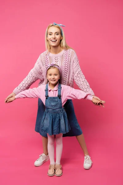 Longitud completa de la madre y el niño sonriendo y de pie con las manos extendidas aisladas en rosa - foto de stock