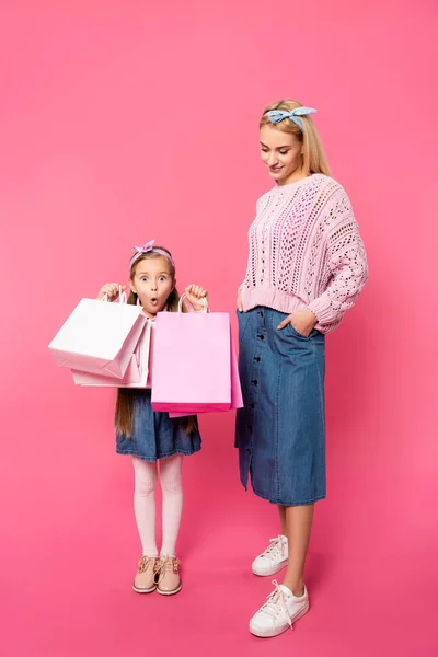 Pleine longueur de mère heureuse debout avec la main dans la poche et regardant fille choquée avec des sacs en papier sur rose — Photo de stock