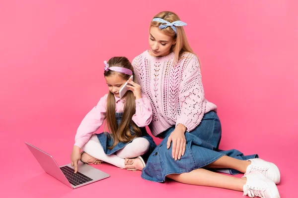 Kid talking on smartphone and using laptop while sitting with mother on pink — Stock Photo