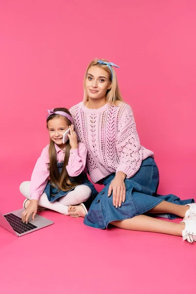 Niño feliz hablando en el teléfono inteligente y el uso de la computadora portátil mientras está sentado con la madre en rosa - foto de stock