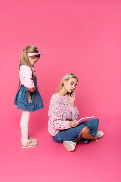 Ofendido niño mirando a la madre hablando en el teléfono inteligente y el uso de la tableta digital en rosa - foto de stock