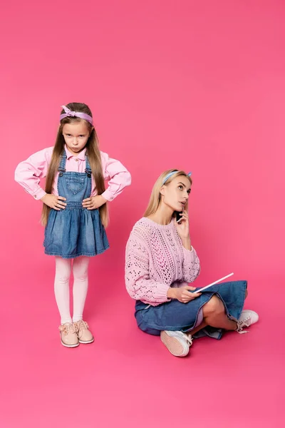 Criança ofendida de pé com as mãos em quadris mãe falando no smartphone e usando tablet digital em rosa — Fotografia de Stock