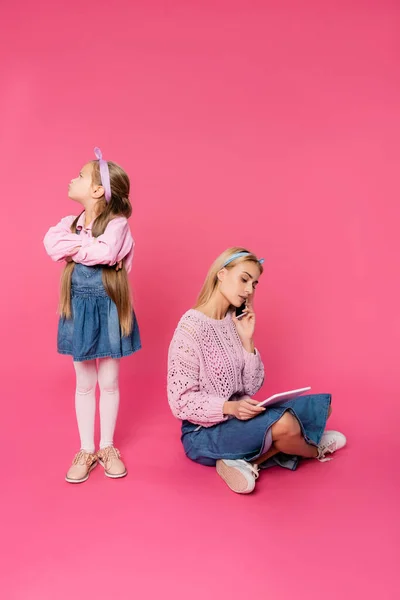 Niño ofendido mirando hacia otro lado mientras estaba de pie con los brazos cruzados cerca de la madre hablando en el teléfono inteligente y utilizando la tableta digital en rosa - foto de stock