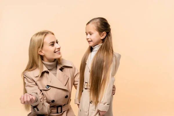 Madre e hija en abrigos de trinchera mirándose y sonriendo aisladas en beige - foto de stock