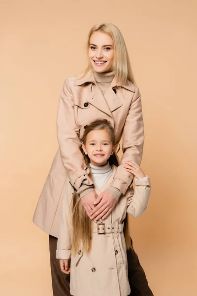 Mutter und Tochter in Trenchcoats umarmen und lächeln isoliert auf beige — Stockfoto