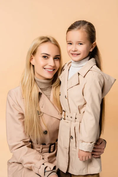 Joyful mother and daughter in trench coats looking at camera isolated on beige — Stock Photo