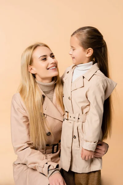 Alegre madre e hija en abrigos de trinchera mirándose y sonriendo aisladas en beige - foto de stock