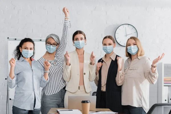 Excited multicultural businesswomen in medical masks near desk — Stock Photo