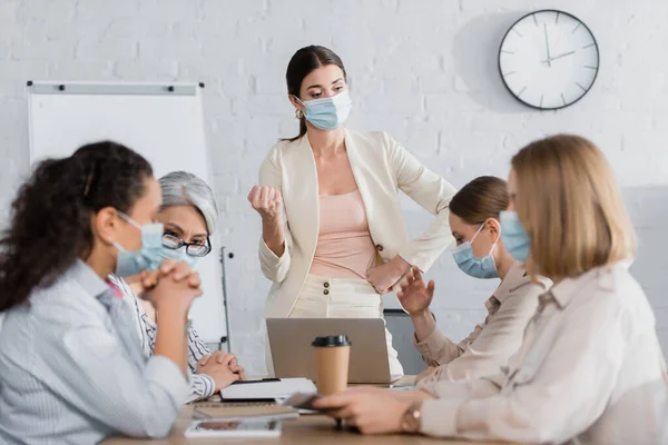 Líder de equipo en máscara médica cerca de compañeros de trabajo multiculturales durante la reunión - foto de stock