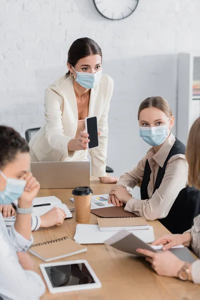 Лидер команды в медицинской маске держит смартфон с чистым экраном рядом с межрасовыми деловыми женщинами — стоковое фото