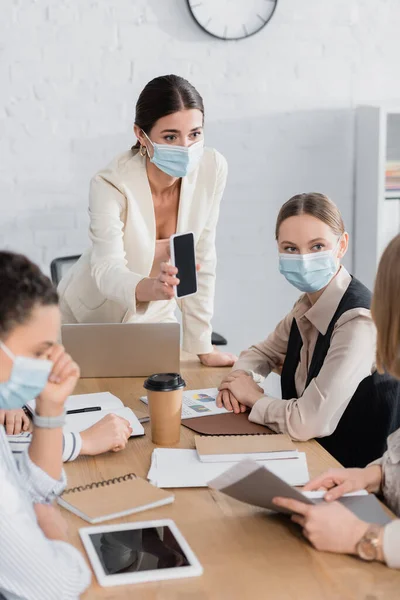 Leader del team in mascherina medica in possesso di smartphone con schermo bianco vicino a donne d'affari interrazziale — Foto stock