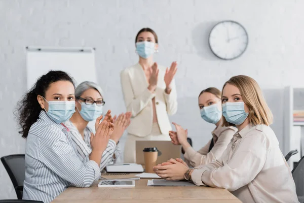 Interracial businesswomen in medical masks applauding with team leader on blurred background — Stock Photo