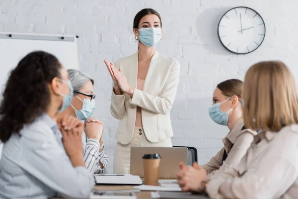 Junge Teamleiterin in medizinischer Maske sieht interrassische Geschäftsfrauen im verschwommenen Vordergrund — Stockfoto