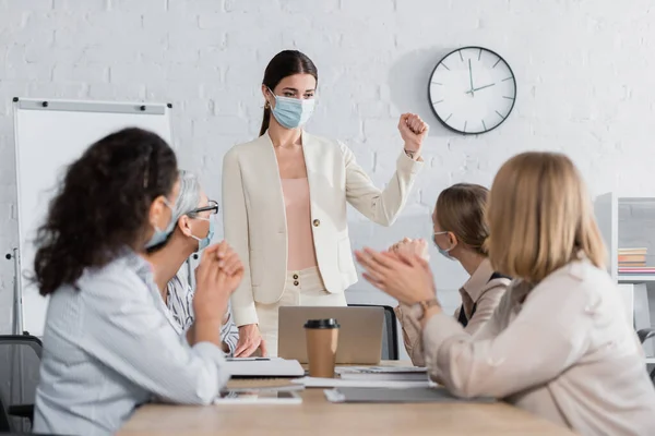 Young team leader in medical mask standing with clenched fist interracial businesswomen on blurred foreground — Stock Photo