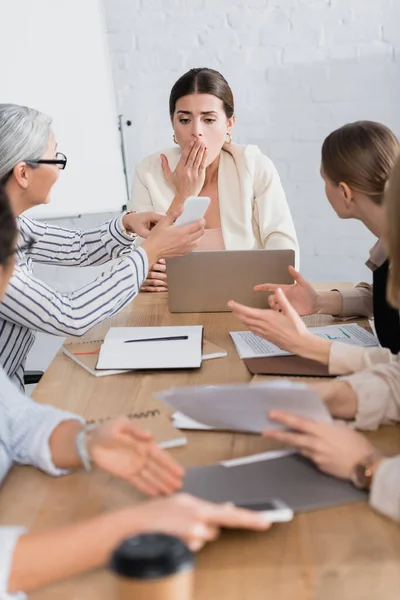 Shocked businesswoman covering mouth and looking at smartphone near interracial colleagues on blurred foreground — Stock Photo