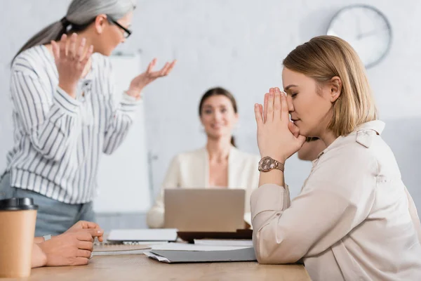 Besorgte Geschäftsfrau mit geschlossenen Augen und betenden Händen am Schreibtisch mit interrassischen Kollegen auf verschwommenem Hintergrund — Stockfoto