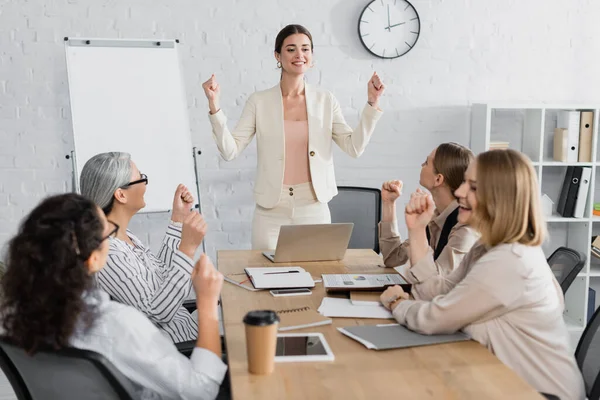 Sonriente líder del equipo de pie con los puños cerrados cerca de las empresarias multiculturales en primer plano borroso - foto de stock