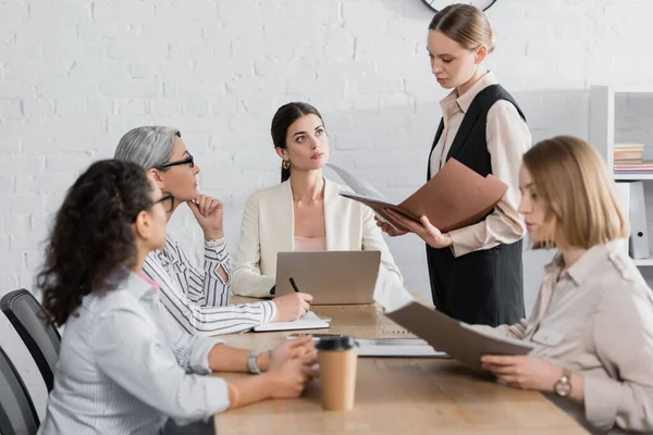 Joven empresaria sosteniendo carpeta cerca del líder del equipo y compañeros de trabajo interracial durante la reunión - foto de stock