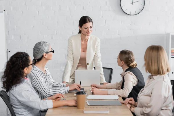 Heureux chef d'équipe debout près de femmes d'affaires multiethniques lors d'une réunion au bureau — Photo de stock