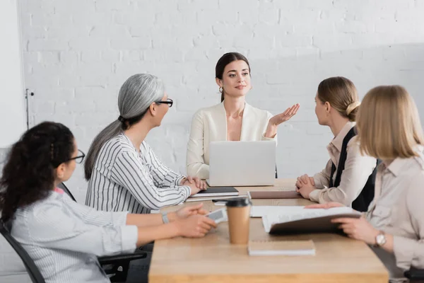 Happy team leader pointing with hand near laptop and multicultural businesswomen during meeting — Stock Photo