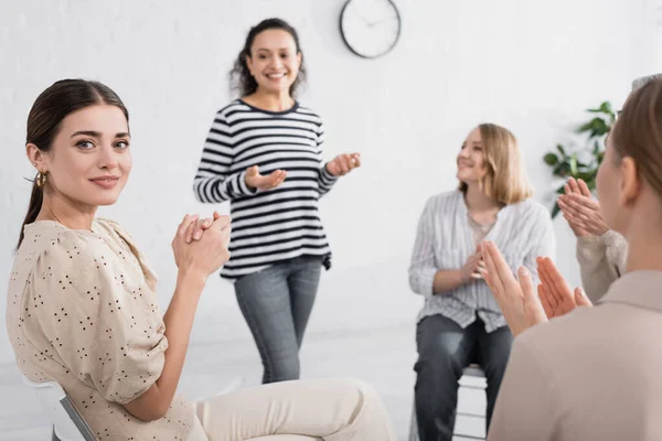 Joven mujer aplaudiendo y mirando a la cámara cerca de afroamericano altavoz de pie sobre fondo borroso - foto de stock