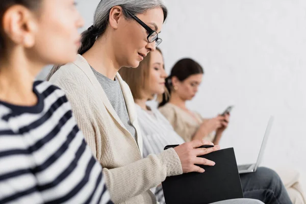 Asian businesswoman in glasses holding pen and notebook near coworkers during lecture — Stock Photo