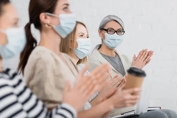 Asiático mujer de negocios en médico máscara aplaudiendo con colegas durante seminario - foto de stock