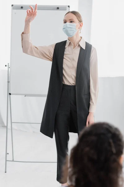 Speaker in medical mask raising hand while standing near flipchart — Stock Photo