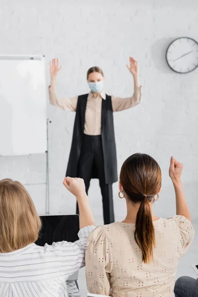Vue arrière des femmes d'affaires levant les mains près du haut-parleur dans le masque médical debout près de tableau à feuilles sur fond flou — Photo de stock