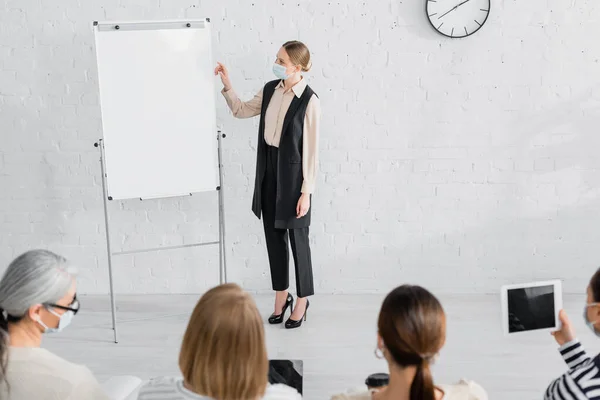 Altoparlante in maschera medica in piedi vicino flipchart e donne d'affari in primo piano sfocato — Foto stock