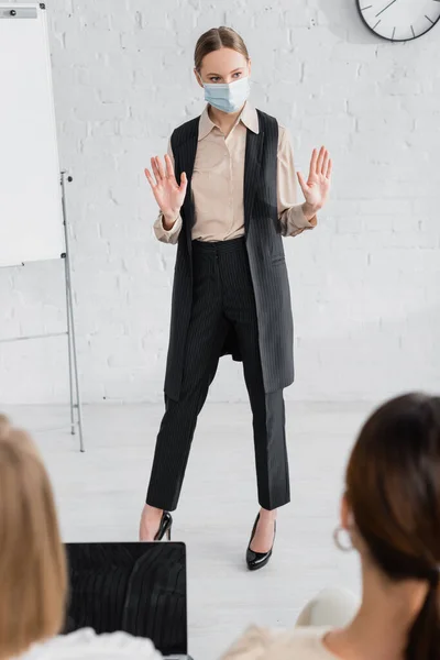 Speaker in medical mask standing and gesturing near flipchart and businesswomen on blurred foreground — Stock Photo