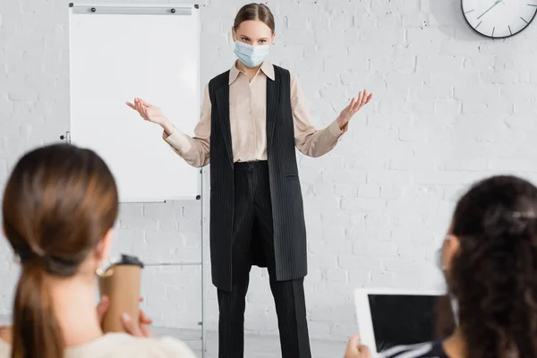 Alto-falante em pé máscara médica e gestos enquanto olha para mulheres de negócios em primeiro plano borrado — Fotografia de Stock