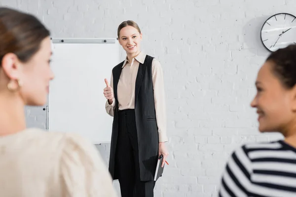 Fröhliche Rednerin zeigt Daumen hoch und sieht interrassische Geschäftsfrauen im verschwommenen Vordergrund an — Stockfoto