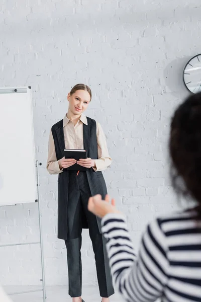 Smiling speaker standing with notebook and looking at businesswoman on blurred foreground during lecture — Stock Photo