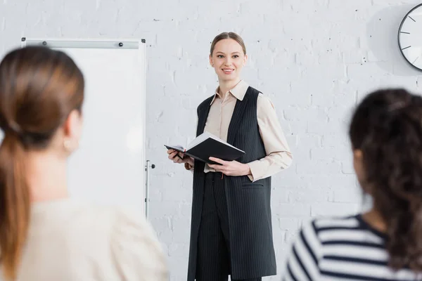 Fröhlicher Redner hält Notizbuch in der Hand und lächelt nahe Frau während Seminar über verschwommenen Vordergrund — Stockfoto