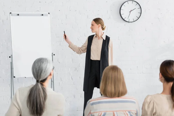 Cheerful speaker looking at blank flipchart near woman during seminar on blurred foreground — Stock Photo